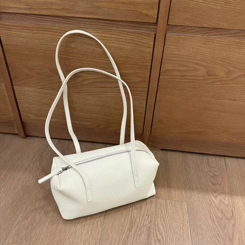 May include: A cream-colored handbag with a rectangular shape and a zipper closure. The bag has two long, thin handles and is made of a textured material. The bag is sitting on a wooden floor.