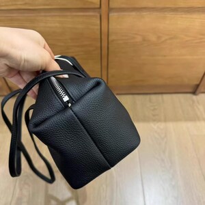 May include: A small, black, textured leather bag with a silver zipper closure. The bag has a rectangular shape with a long, thin strap. The bag is held by a hand, with a wooden cabinet in the background.
