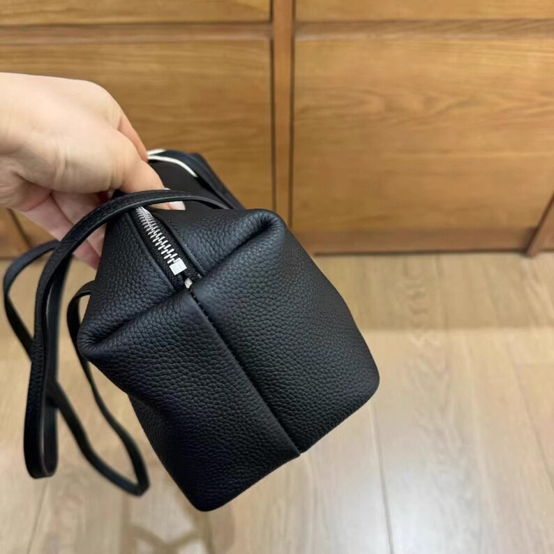 May include: A small, black, textured leather bag with a silver zipper closure. The bag has a rectangular shape with a long, thin strap. The bag is held by a hand, with a wooden cabinet in the background.