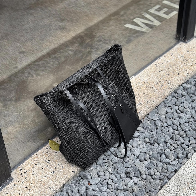 May include: A black woven tote bag with black leather handles and a small matching pouch. The bag is rectangular and has a textured appearance. The bag is sitting on a stone surface.
