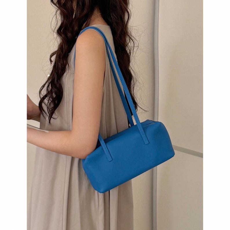 May include: A vibrant blue rectangular handbag with two long straps. The bag is held by a person wearing a neutral-colored sleeveless dress. The bag's design is simple and sleek, with a focus on the bright color and clean lines.