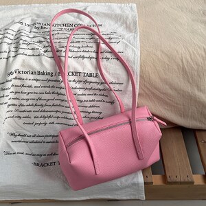May include: A pink rectangular handbag with a zipper closure and long straps. The bag is made of a textured material and is set against a white cloth with printed text.