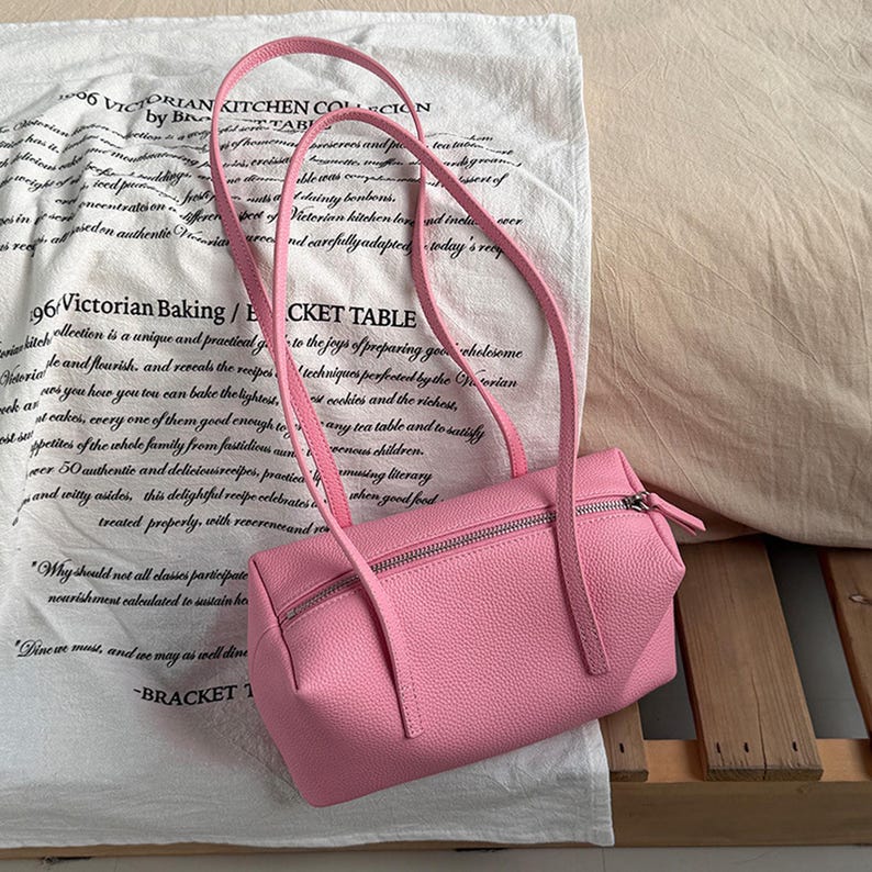 May include: A pink rectangular handbag with a zipper closure and long straps. The bag is made of a textured material and is set against a white cloth with printed text.