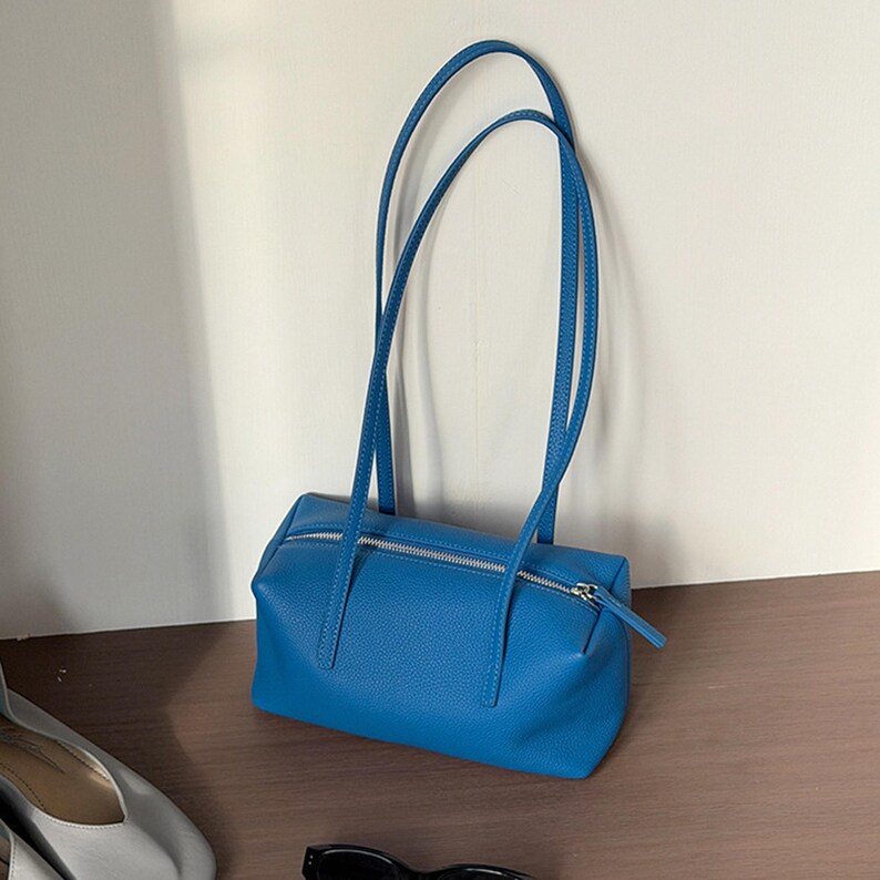May include: A bright blue handbag with long, thin straps. The rectangular bag has a silver zipper closure and is made of a textured material. The bag is sitting on a wooden surface.
