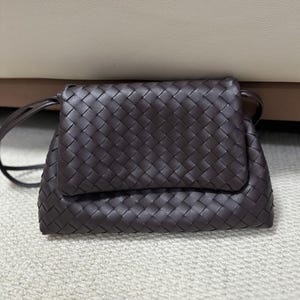 May include: A dark brown, woven leather crossbody bag with a flap closure. The bag features a long, thin strap and a textured, basketweave pattern. The bag is rectangular and appears to be made of high-quality leather.