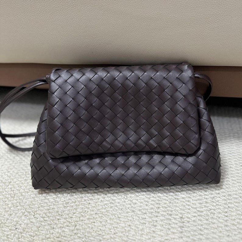May include: A dark brown, woven leather crossbody bag with a flap closure. The bag features a long, thin strap and a textured, basketweave pattern. The bag is rectangular and appears to be made of high-quality leather.