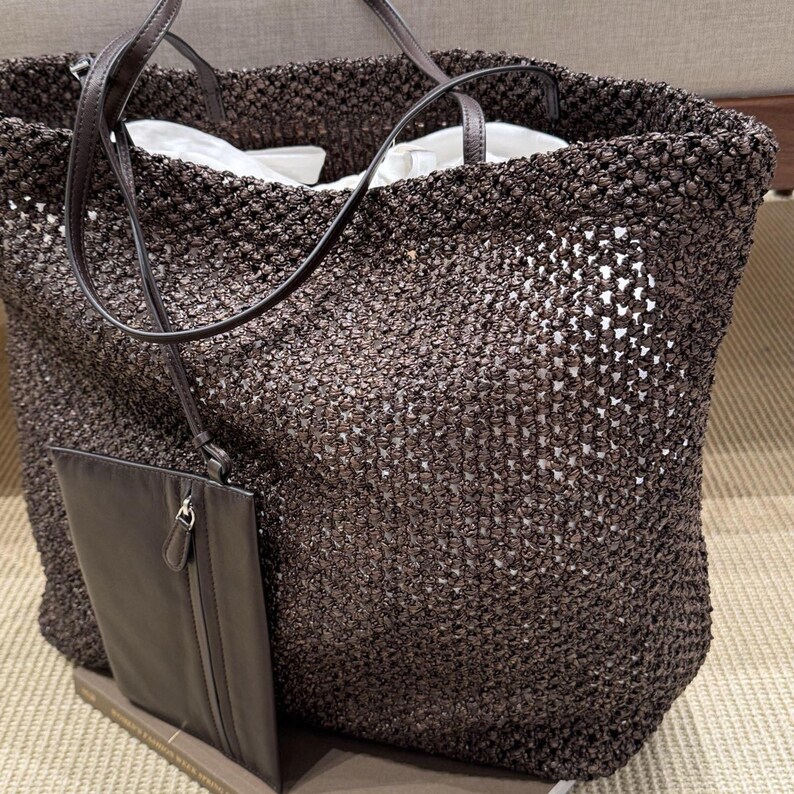 May include: A large, open-weave tote bag in a dark brown color. The bag has a matching dark brown leather strap and a small, attached pouch. The interior of the bag is lined with white fabric. The bag is sitting on a light brown surface.