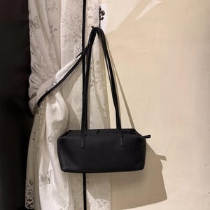 May include: A black rectangular handbag with long, thin straps hangs from a hook. The bag has a textured surface and a simple, minimalist design. The background includes a white curtain with a floral pattern.