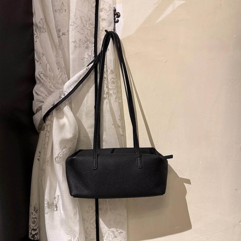 May include: A black rectangular handbag with long, thin straps hangs from a hook. The bag has a textured surface and a simple, minimalist design. The background includes a white curtain with a floral pattern.