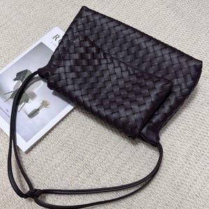 May include: A dark brown woven leather clutch bag with a matching smaller pouch. The bag has a long, thin strap and is resting on a magazine. The woven pattern is consistent across both pieces, creating a cohesive design.
