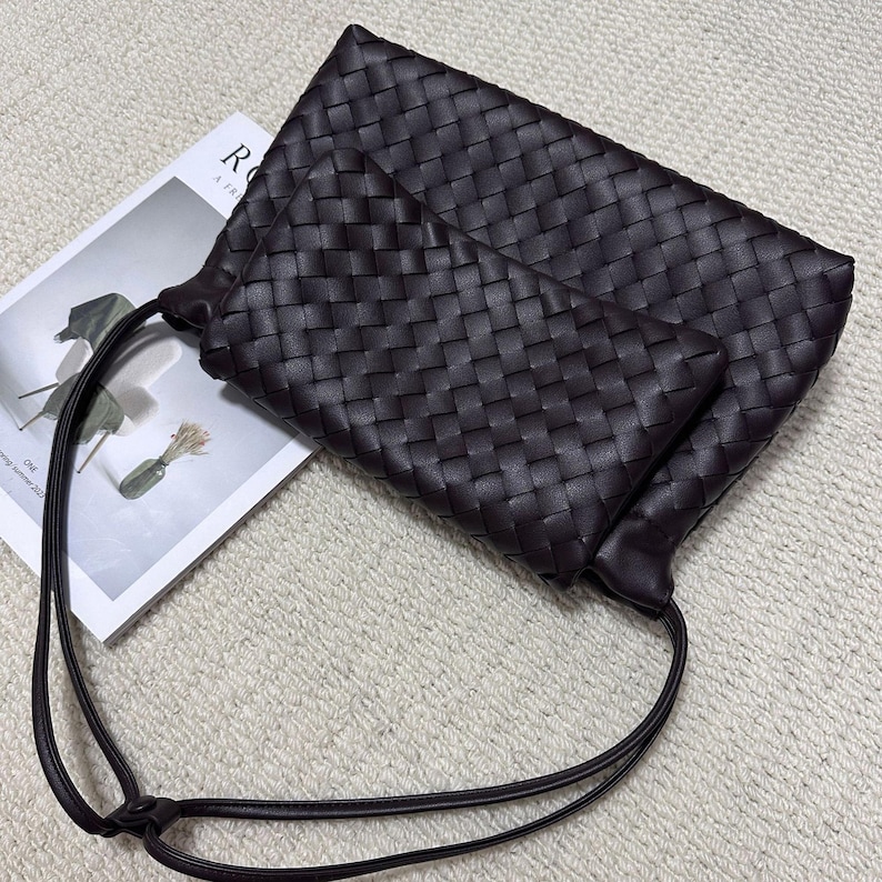 May include: A dark brown woven leather clutch bag with a matching smaller pouch. The bag has a long, thin strap and is resting on a magazine. The woven pattern is consistent across both pieces, creating a cohesive design.