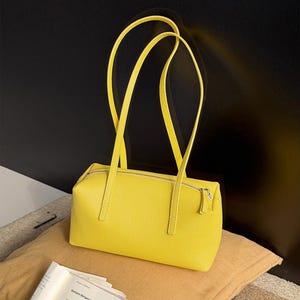 May include: A bright yellow rectangular handbag with long, thin handles. The bag has a zipper closure and is made of textured material. The bag is sitting on a tan pillow with a black background.