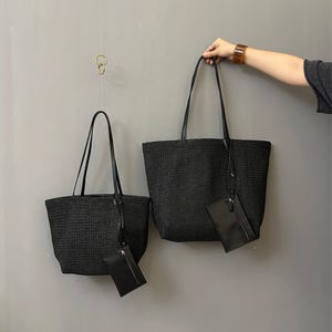 May include: Two black woven tote bags with matching small pouches. The bags have long, black handles and are hanging against a gray background. One bag is held by a person. The bags are of different sizes.