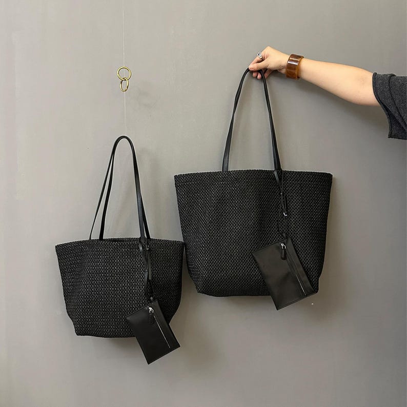 May include: Two black woven tote bags with matching small pouches. The bags have long, black handles and are hanging against a gray background. One bag is held by a person. The bags are of different sizes.