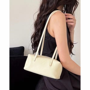 May include: Cream-colored rectangular handbag with long, thin straps. The bag has a textured surface and a zipper closure. The straps are a lighter shade of cream. The bag is held against a black background.