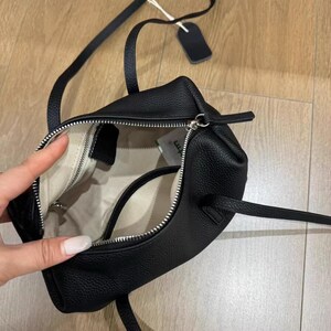 May include: Black leather handbag with a top zipper closure and a long, adjustable strap. The bag has a structured shape with a textured surface. The interior is lined with a light-colored fabric. A small tag hangs from the strap.