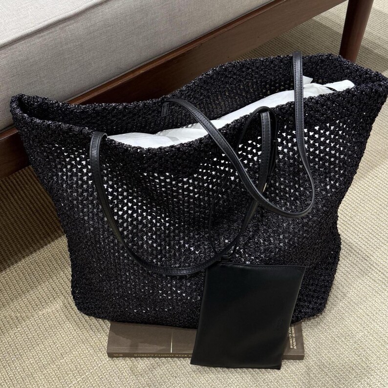 May include: Black woven tote bag with leather handles and a matching small pouch. The open-weave design reveals a white interior. The bag is sitting on a neutral-colored surface.