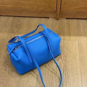 May include: A bright blue handbag with a rectangular shape and a textured surface. The bag features two short handles and a long, adjustable strap. Silver zippers secure the top and side compartments. The bag is sitting on a wooden floor.