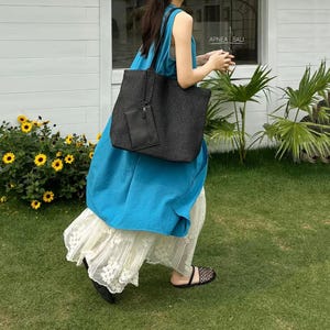 May include: A black woven tote bag with a matching small pouch, carried by a person wearing a blue dress and a white lace skirt. The bag has a rectangular shape and a simple design. The scene is outdoors with green grass and yellow flowers.