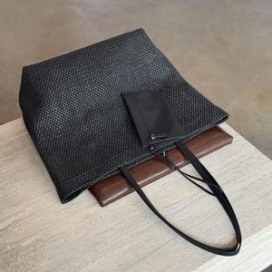 May include: A black woven tote bag with a brown leather base and a small black pouch. The bag has long, thin black straps. The tote bag is sitting on a light-colored surface.
