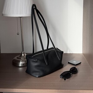 May include: A black leather handbag with long straps, a zippered top, and a rectangular shape. The bag is on a wooden surface next to a pair of sunglasses and a small compact. A silver lamp with a white shade is in the background.