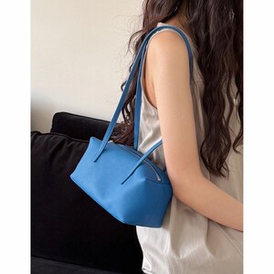 May include: A bright blue rectangular handbag with a zipper closure and long straps. The bag has a textured surface and is being carried over the shoulder. The bag is a vibrant shade of blue.