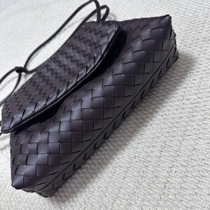 May include: A dark brown leather crossbody bag with a woven pattern. The bag has a rectangular shape with a flap closure and a long, thin strap. The leather appears to be soft and supple.