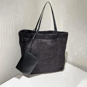 May include: A black woven tote bag with long, thin straps and a matching small pouch. The bag has a rectangular shape and a textured, open weave design. The pouch is smooth and rectangular.