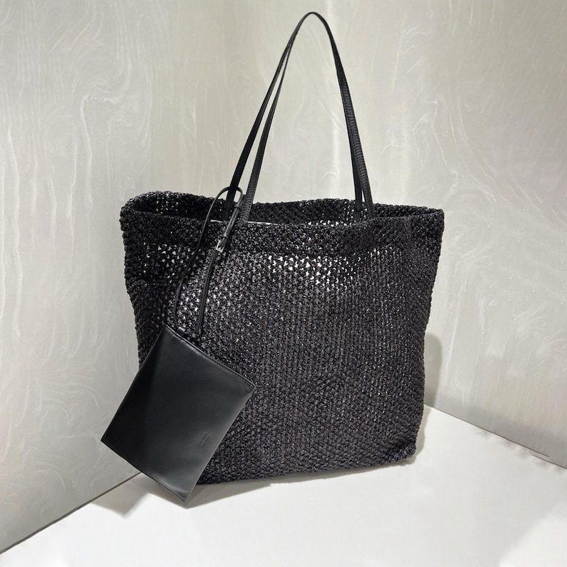 May include: A black woven tote bag with long, thin straps and a matching small pouch. The bag has a rectangular shape and a textured, open weave design. The pouch is smooth and rectangular.