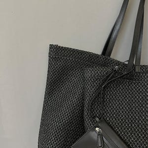 May include: A black woven tote bag with black leather handles and a small black wallet. The tote bag has a textured, open weave design. The wallet has a zipper closure and a small strap.