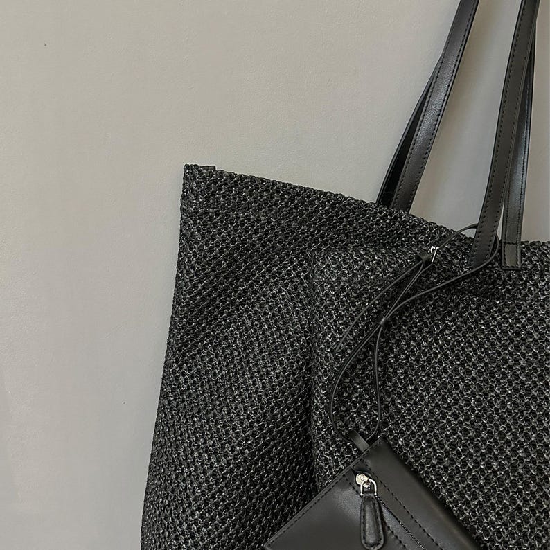 May include: A black woven tote bag with black leather handles and a small black wallet. The tote bag has a textured, open weave design. The wallet has a zipper closure and a small strap.