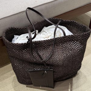 May include: A dark brown woven tote bag with a matching small pouch attached. The bag has long, thin handles and is filled with white fabric. The bag is sitting on a light-colored rug.
