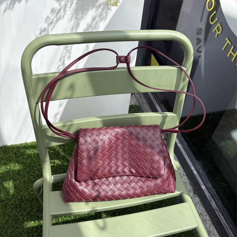 May include: A burgundy leather crossbody bag with a woven design, resting on a light green chair. The bag features a long, thin strap and a flap closure. The bag's color contrasts with the chair, creating a visually appealing composition.