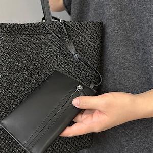 May include: Black woven tote bag with a matching black leather wallet. The wallet has a zipper closure and a wrist strap. The tote bag has a long strap. The bag and wallet are held by a person.