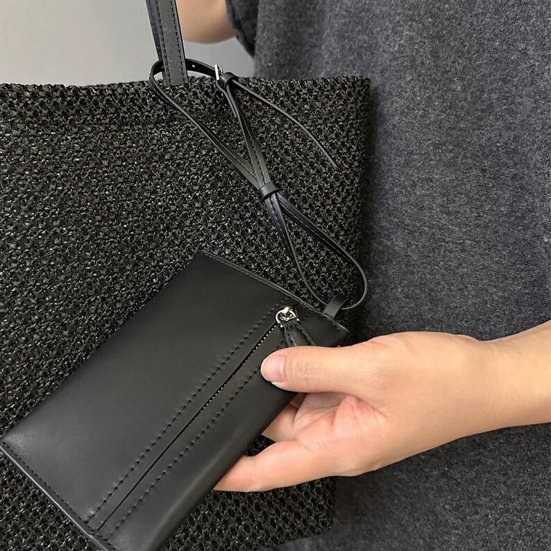 May include: Black woven tote bag with a matching black leather wallet. The wallet has a zipper closure and a wrist strap. The tote bag has a long strap. The bag and wallet are held by a person.