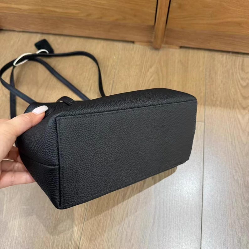 May include: A black rectangular handbag with a textured surface. The bag has a long strap and a small handle. The bag is held by a hand with manicured nails. The bag is sitting on a wooden surface.