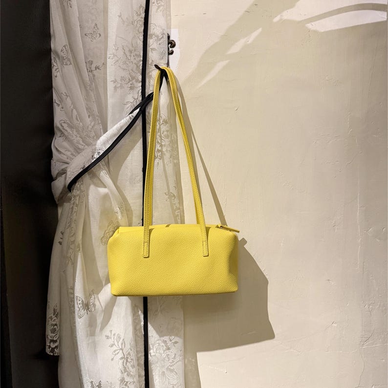 May include: A bright yellow rectangular handbag with long, thin straps hangs from a hook. The bag has a textured surface and a zipper closure. The background includes a white curtain with a floral pattern.