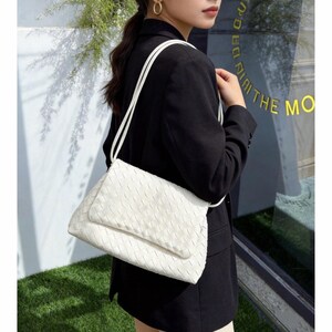 May include: A white woven shoulder bag with a flap closure and long, thin straps. The bag is rectangular and appears to be made of a soft material. The background includes greenery and a window with text.