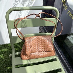 May include: A brown woven leather crossbody bag with a flap closure and a long, thin strap. The bag is resting on a light green metal chair, with the strap draped over the chair's back. The bag's woven texture is visible.