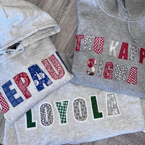 May include: Three gray hooded sweatshirts with collegiate lettering. The sweatshirts feature the names "DePaul", "Tau Kappa Sigma", and "Loyola" in various patterned fabrics, including paisley, gingham, and floral designs. The sweatshirts are folded.