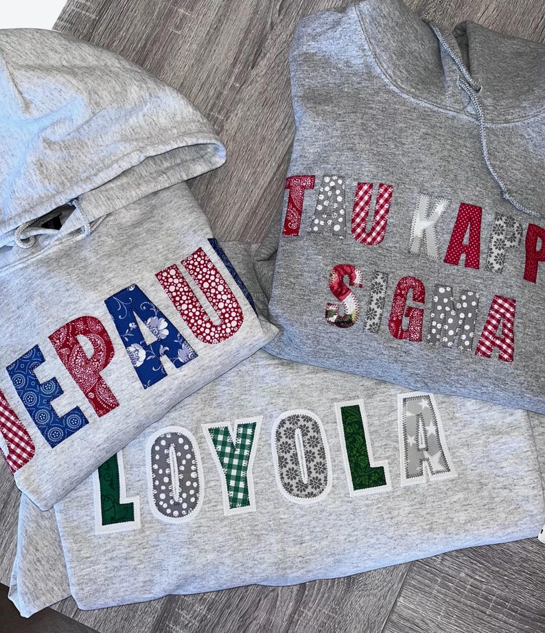 May include: Three gray hooded sweatshirts with collegiate lettering. The sweatshirts feature the names "DePaul", "Tau Kappa Sigma", and "Loyola" in various patterned fabrics, including paisley, gingham, and floral designs. The sweatshirts are folded.