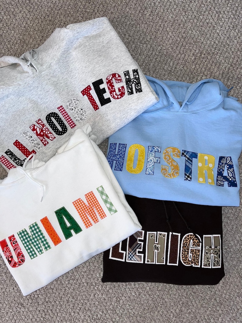 May include: Four hooded sweatshirts in gray, white, light blue, and brown. Each sweatshirt features a university name in colorful, patterned letters: Illinois Tech, U Miami, Hofstra, and Lehigh. The sweatshirts are folded and stacked on a textured surface.