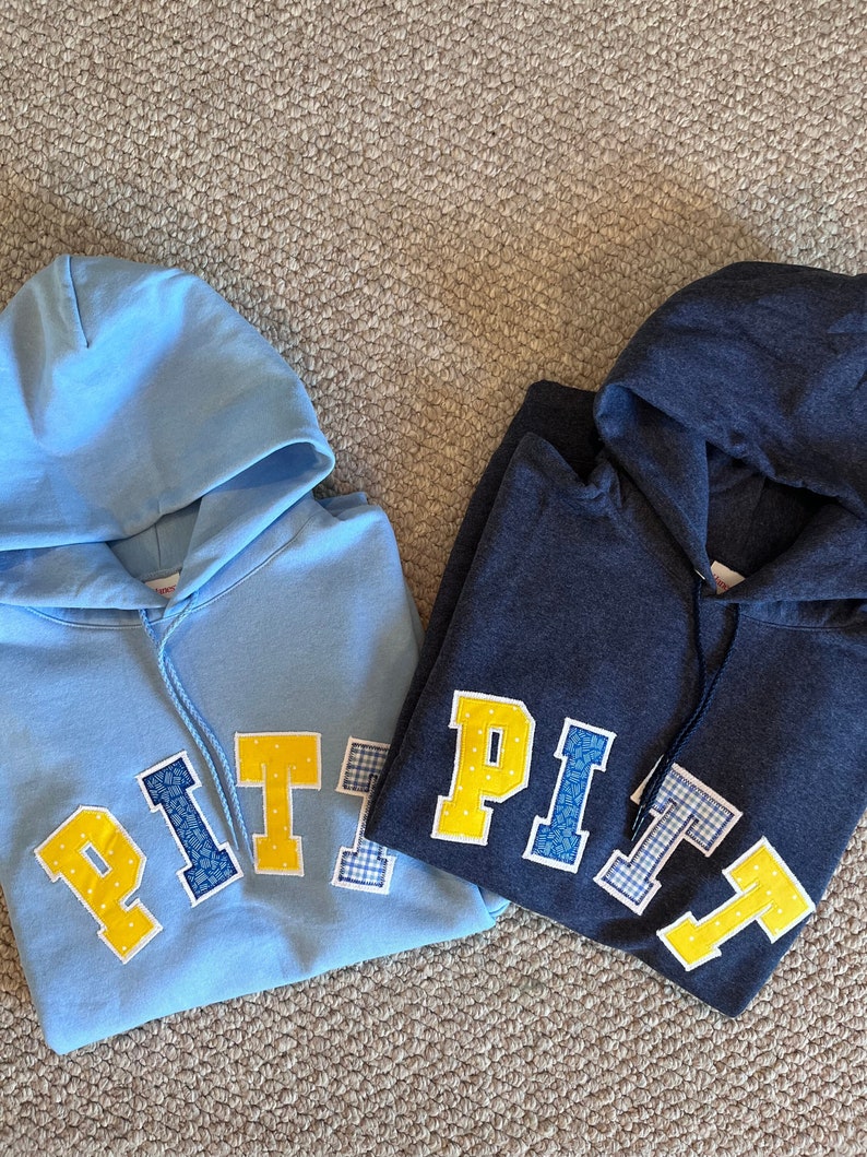 May include: Two hooded sweatshirts, one light blue and one navy blue, are displayed. Each features the word "PITT" in a collegiate-style font. The letters are yellow with white polka dots, blue floral, and blue and white gingham patterns.