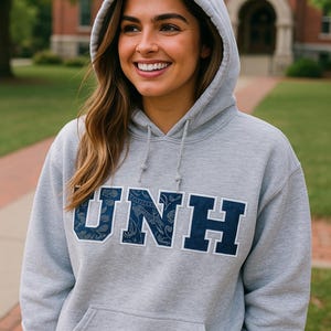 May include: A gray hooded sweatshirt with the letters "UNH" in navy blue, with a paisley pattern within the letters. The sweatshirt has a front pocket and a drawstring hood. The person is smiling.