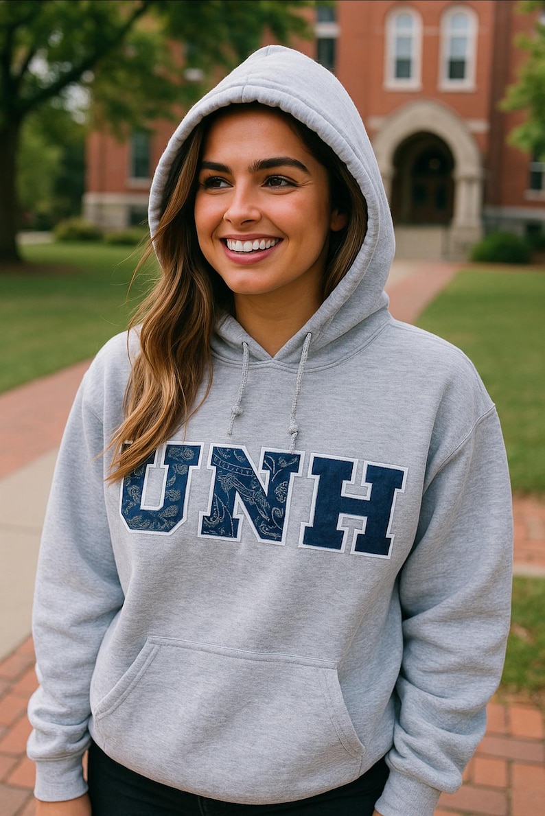 May include: A gray hooded sweatshirt with the letters "UNH" in navy blue, with a paisley pattern within the letters. The sweatshirt has a front pocket and a drawstring hood. The person is smiling.
