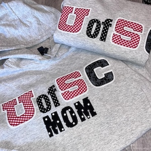 May include: Two gray hoodies with the text "U of SC" in red gingham and black polka dot letters. One hoodie also says "MOM" in black polka dot letters. The hoodies are on a wooden surface.