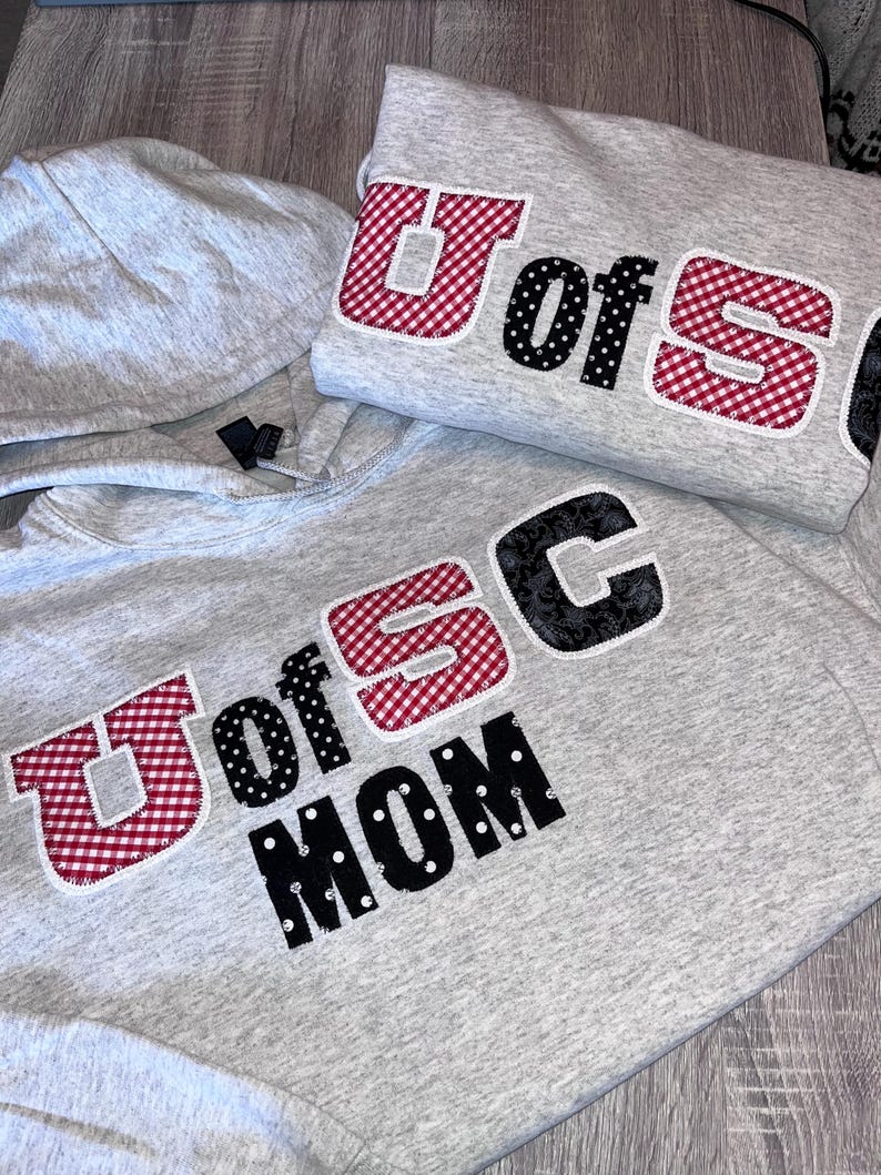 May include: Two gray hoodies with the text "U of SC" in red gingham and black polka dot letters. One hoodie also says "MOM" in black polka dot letters. The hoodies are on a wooden surface.