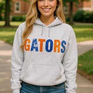 May include: A light gray hoodie with the word "GATORS" in orange and blue lettering. The hoodie has a front pocket and a drawstring hood. The person is wearing denim shorts.