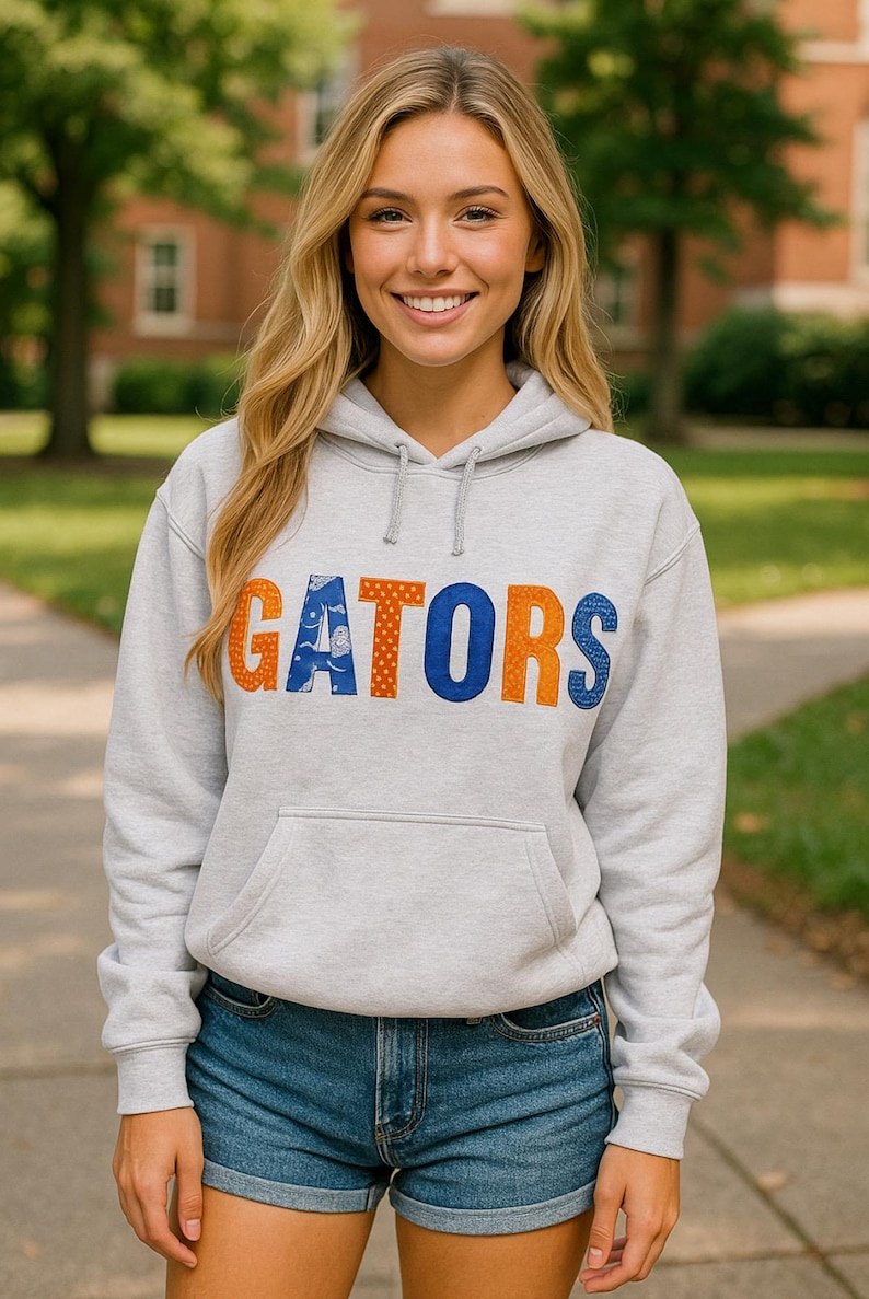 May include: A light gray hoodie with the word "GATORS" in orange and blue lettering. The hoodie has a front pocket and a drawstring hood. The person is wearing denim shorts.