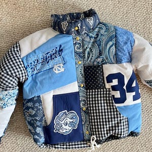 May include: A patchwork puffer jacket featuring various blue, white, and black fabrics. It includes panels with the text North Carolina, the number 34, a ram mascot logo, paisley patterns, gingham checks, and floral prints with a snap button closure.